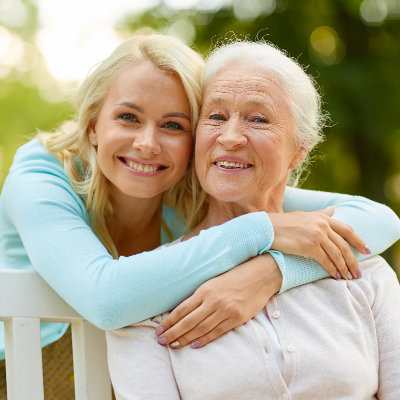 A young woman with blond hair hugs an older woman with gray hair, both smiling and seated on a bench outdoors.