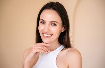 A woman with long brown hair wearing a white sleeveless top smiles at the camera, her hand resting near her chin.