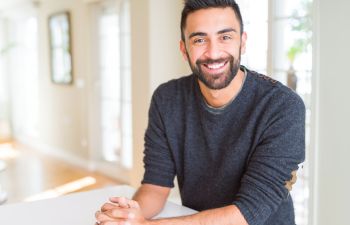 A bearded man with dark hair wearing a grey sweater is smiling at the camera while seated indoors in a bright room.