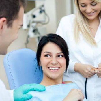 A woman in a dental chair smiles at a dentist, while a hygienist prepares tools in the background.