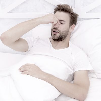 A man in a white shirt lies in bed, holding a pillow and rubbing his eyes.