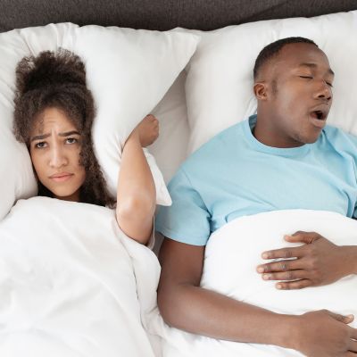 A woman covers her ear with a pillow while a man sleeps next to her, appearing to snore.