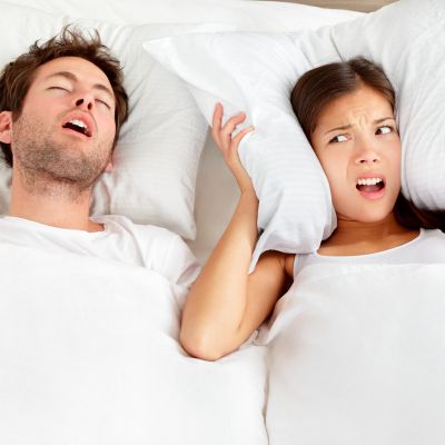 A man is sleeping and snoring loudly. A woman lying next to him looks annoyed and holds a pillow over her ear.