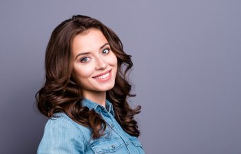 A beautiful young woman with long, wavy brown hair and light-colored eyes smiles warmly as she looks over her shoulder at the camera. She is wearing a blue denim shirt and is set against a plain gray background with ample copy space to the right.