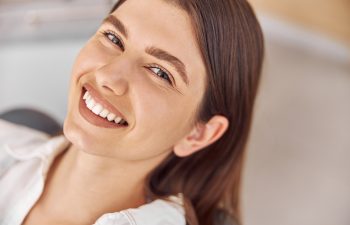 A close-up, slightly high-angle shot of a smiling woman with long brown hair, wearing a white collared shirt, showing off her teeth. The background is soft and brightly lit, suggesting an indoor setting like a dentist's office.