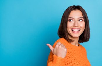A cheerful young woman in an orange sweater smiling and pointing her thumb to the side against a blue background.