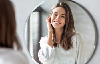 A smiling young woman with brown hair, wearing a white silk-like robe, looks at her reflection in a round bathroom mirror and gently touches her hair.