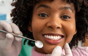 A close-up of a young woman smiling at the dentist, while gloved hands hold a dental mirror and gently touch her chin.