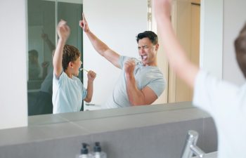 A man and a young child dance and sing energetically in front of a bathroom mirror, while the child holds a toothbrush.