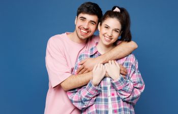 Young man in a pink shirt hugs a smiling young woman in a plaid shirt, both standing in front of a blue background.