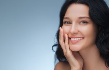 A portrait of a woman with dark wavy hair smiling and gently touching her cheek, showcasing her white teeth against a soft blue background.