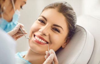 Close-up of a smiling young woman lying in a dental chair while a dentist in blue gloves examines her white teeth with metal tools.